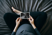 High angle shot of Asian pregnant woman holding an ultrasound scan photo in front of her baby bump, sitting on bed at home. Mother-to-be. Precious moment in life. Preparation for a new family member. Expecting a new life. Baby and new life concep
