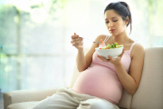 Cute pregnant brunete woman relaxing on sofa and enjoying a vegetable salad.She's in late 20's.Wearing beige pregnancy pants and pink sleeveless tank to