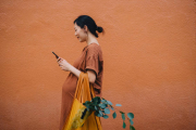 Young Asian pregnant woman carrying a yellow reusable shopping bag and using smartphone, shopping for fresh groceries in the city. Standing against orange wall. Responsible shopping, zero waste, sustainable and healthy eating lifestyle during pregnanc