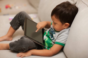 Comfortably sitting on a sofa with a tablet on his lap, a little boy uses his index finger to turn on the device