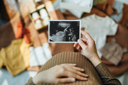 Personal perspective of Asian pregnant woman looking at an ultrasound scan photo while gently touching her baby bump, with baby clothings and accessories laying on the floor. Mother-to-be. Preparation for a new family member. Expecting a new life concep