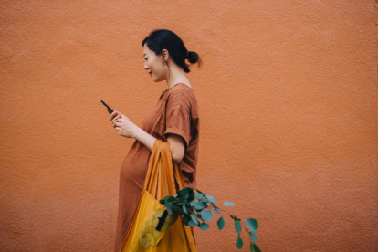 Young Asian pregnant woman carrying a yellow reusable shopping bag and using smartphone, shopping for fresh groceries in the city. Standing against orange wall. Responsible shopping, zero waste, sustainable and healthy eating lifestyle during pregnanc