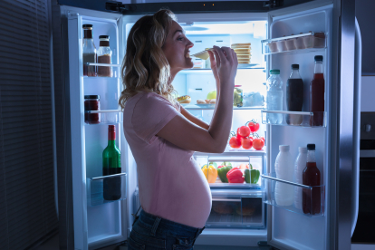Close-up Of A Young Pregnant Woman Eating Slice Of Cheese In Front Of Refrigerator