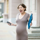 Portrait of unhappy young pregnant business woman walking in the city with document folder and mobile phone. Irritated office worker pregnant woman using smartphone on the stree