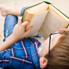 Little boy is reading a book while laying on bed and wearing glasse