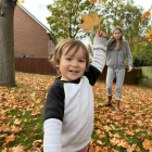 A young boy is standing on an autumn lawn of fallen leaves. He is holding his left arm in the air with a large golden leaf in his hand. He is smiling at the camera with his cousin in the background at a distance behind. Houses and deciduous trees are in the background.