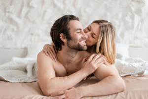 Affectionate young couple lying together in bed, hugging and kissing after lovemaking. Cheerful millennial lovers relaxing and cuddling after having sex. Intimate relationship concept