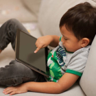 Comfortably sitting on a sofa with a tablet on his lap, a little boy uses his index finger to turn on the device